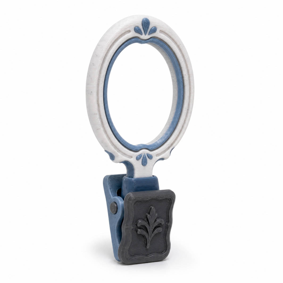 Product illustration of "oval curtain ring with ornamental clip" on white background: curtain ring; Basic shape: large oval ring shape with attached rectangular clip; Design: a double bordered ring edge and a reliefed clip field; Colours: white, grey and dark grey.